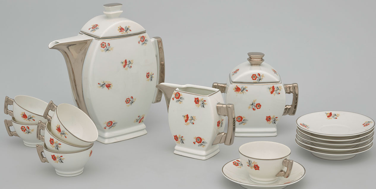 Porcelanowy komplet do kawy na jasnoszarym tle — kremowy dzbanek z pokrywką, mlecznik i cukiernica oraz filiżanki ze spodkami i stos talerzy, wszystkie z drobnymi czerwono-pomarańczowymi motywami kwiatowymi i złotawymi, geometrycznymi uchwytami.