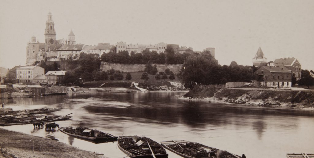 Sepiowe panoramiczne zdjęcie rzeki z kilkoma drewnianymi łodziami załadowanymi ładunkiem przy brzegu, kilkoma zwierzętami stojącymi w płytkiej wodzie po lewej, po drugiej stronie rozległe zadrzewione wzgórze z murem obronnym i zespołem budynków z wysoką wieżą kościelną oraz domami i wieżą po prawej, z odbiciami budowli na spokojnej powierzchni wody.