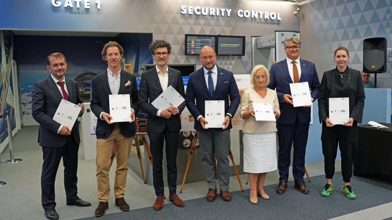 Siedmioro dorosłych stojących w rzędzie przed stanowiskiem oznaczonym „Security Control” i „Gate 1” na lotnisku, każdy trzyma biały folder z logo „I ♥ CEL”, większość w marynarkach i garniturach, jedna kobieta w jasnej spódnicy, druga w czarnym stroju i zielono-żółtych tenisówkach, w tle tablice informacyjne, bramka kontroli bezpieczeństwa i wzorzysta ściana.