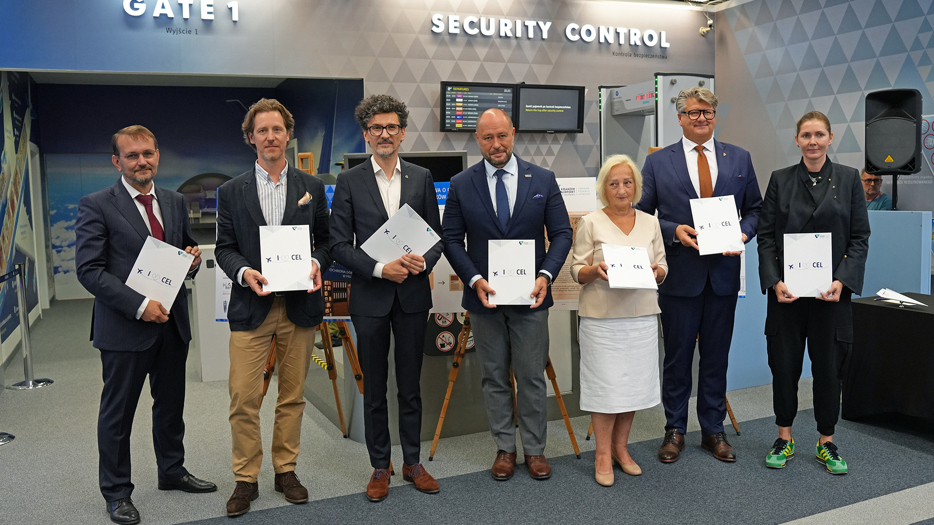 Siedmioro dorosłych stojących w rzędzie przed stanowiskiem oznaczonym „Security Control” i „Gate 1” na lotnisku, każdy trzyma biały folder z logo „I ♥ CEL”, większość w marynarkach i garniturach, jedna kobieta w jasnej spódnicy, druga w czarnym stroju i zielono-żółtych tenisówkach, w tle tablice informacyjne, bramka kontroli bezpieczeństwa i wzorzysta ściana.