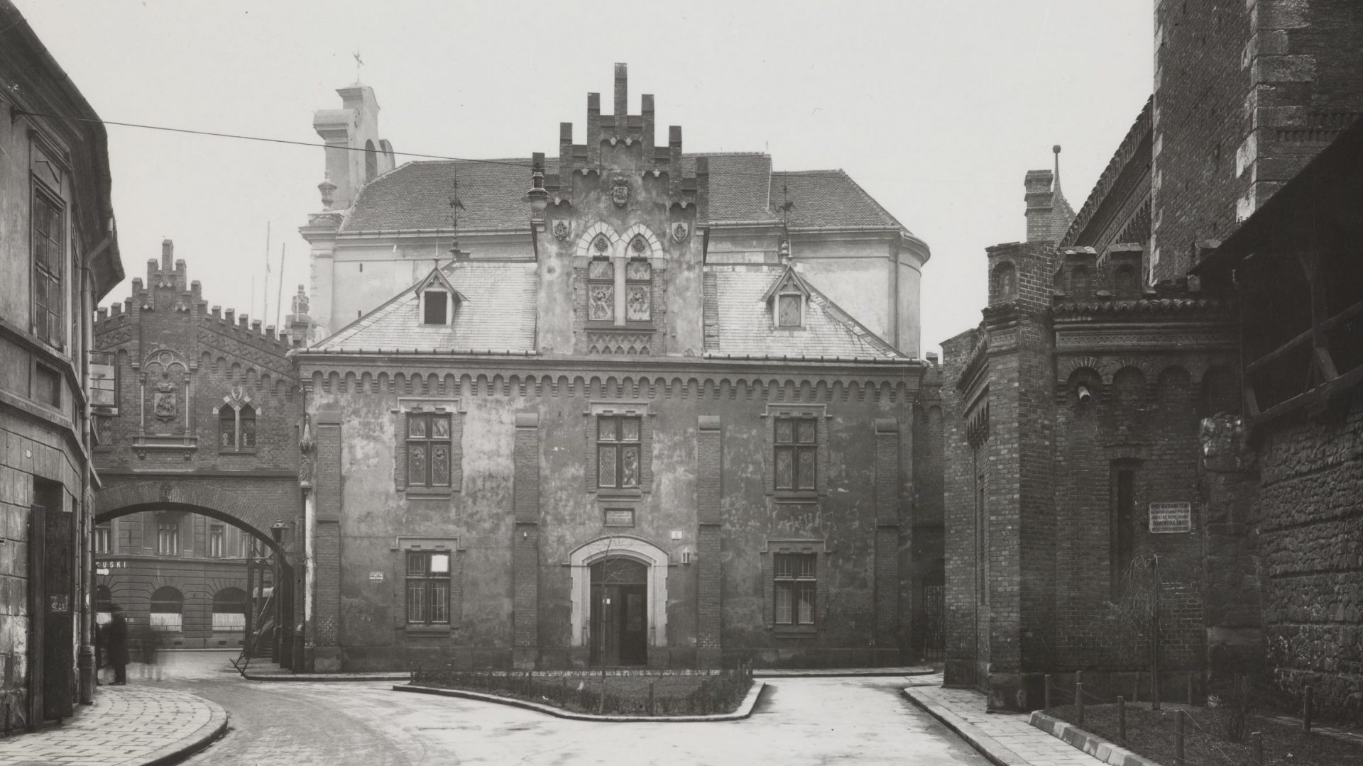 Czarno-białe zdjęcie ukazujące wąską ulicę i frontowy historyczny budynek z ozdobnym schodkowym szczytem, dachami z dwoma lukarnami, kamienno-ceglanymi elewacjami z dekoracyjnymi oknami i tarczami herbów, przed nim mały trójkątny skwer; po lewej przejście pod arkadą do innej kamienicy, po prawej wysoka ceglana ściana z detalami architektonicznymi, a przy łuku kilka rozmytych sylwetek ludzi.
