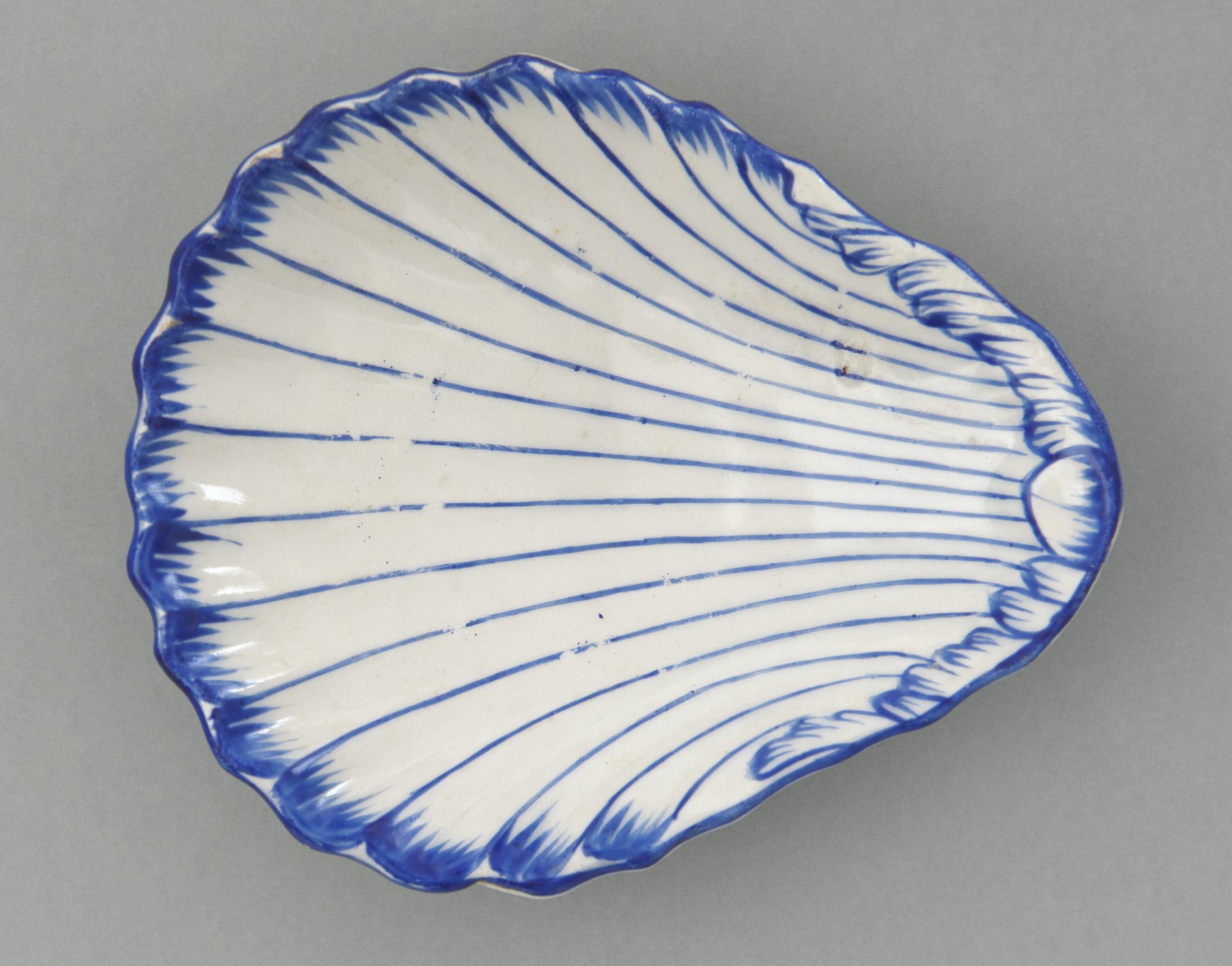 Fragment ceramicznego talerza w kształcie muszli, biały z cienkimi niebieskimi promienistymi liniami i falistym kobaltowym obrzeżem na szarym tle.