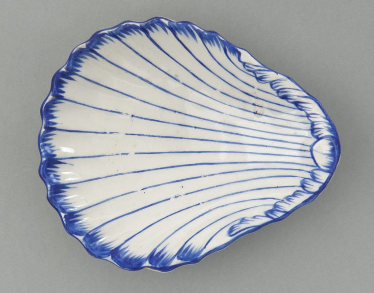 Fragment ceramicznego talerza w kształcie muszli, biały z cienkimi niebieskimi promienistymi liniami i falistym kobaltowym obrzeżem na szarym tle.