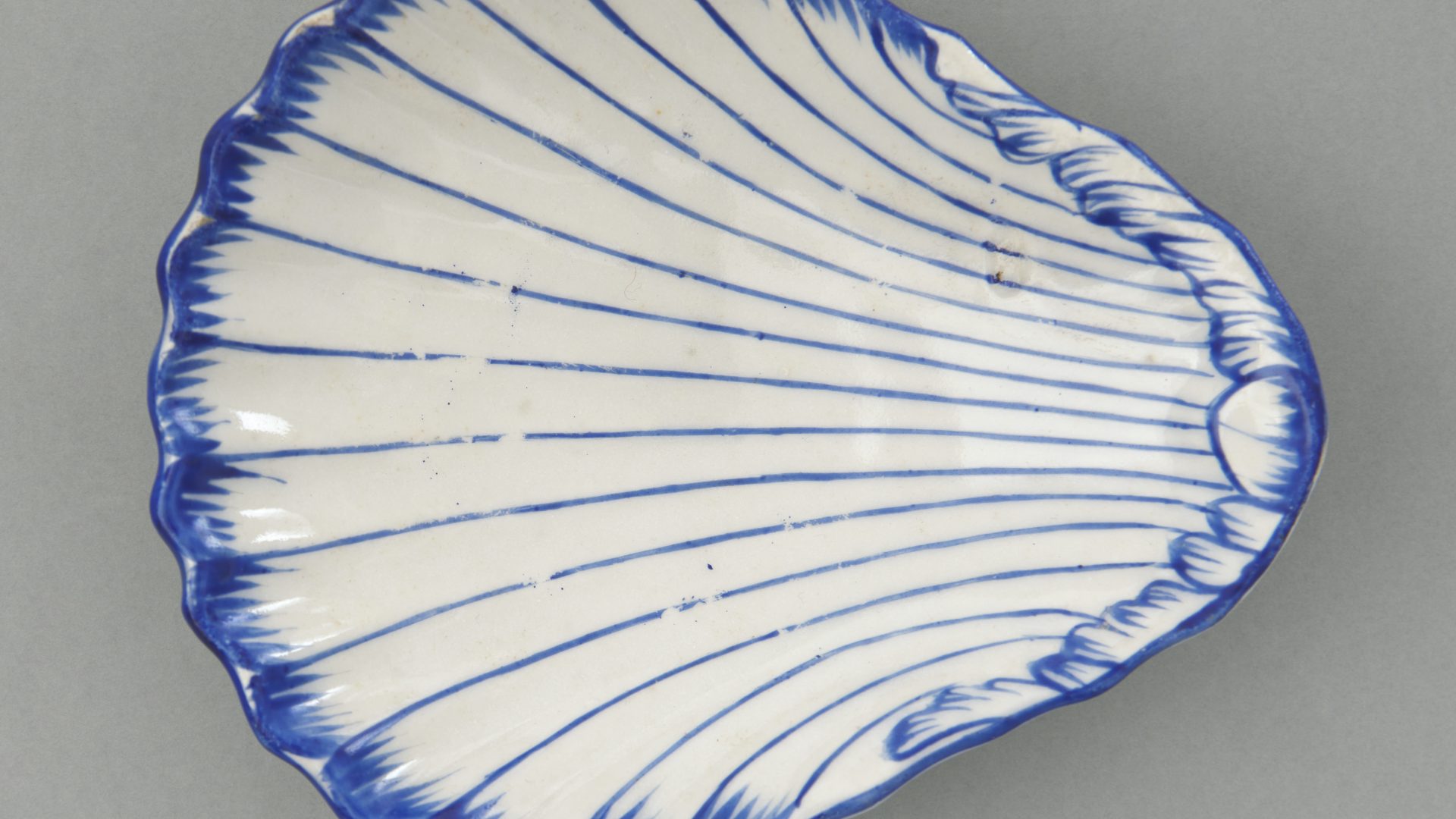 Fragment ceramicznego półmiska w kształcie muszli, biały z promienistymi cienkimi niebieskimi liniami i falistym ciemnoniebieskim brzegiem na szarym tle.