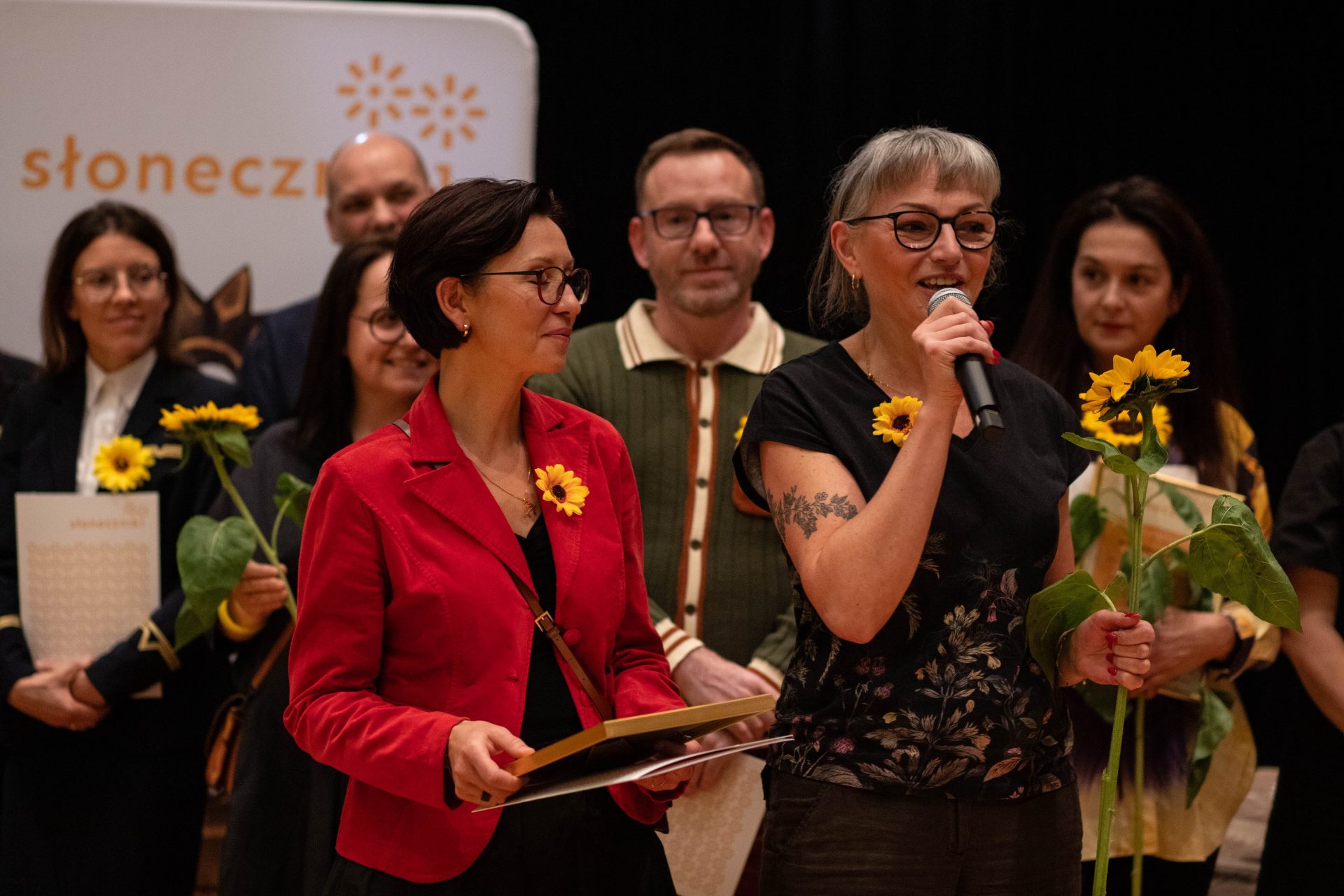Grupa osób stojąca na scenie przed ciemnym tłem; na pierwszym planie po prawej kobieta w okularach z krótkimi siwymi włosami trzyma mikrofon przy ustach i łodygę słonecznika, ma widoczny tatuaż na przedramieniu, po lewej kobieta w czerwonym żakiecie i okularach trzyma oprawiony dyplom i ma przypięty słonecznik, w tle kilka osób trzymających słoneczniki i częściowo widoczny baner z napisem „słonecz…”.