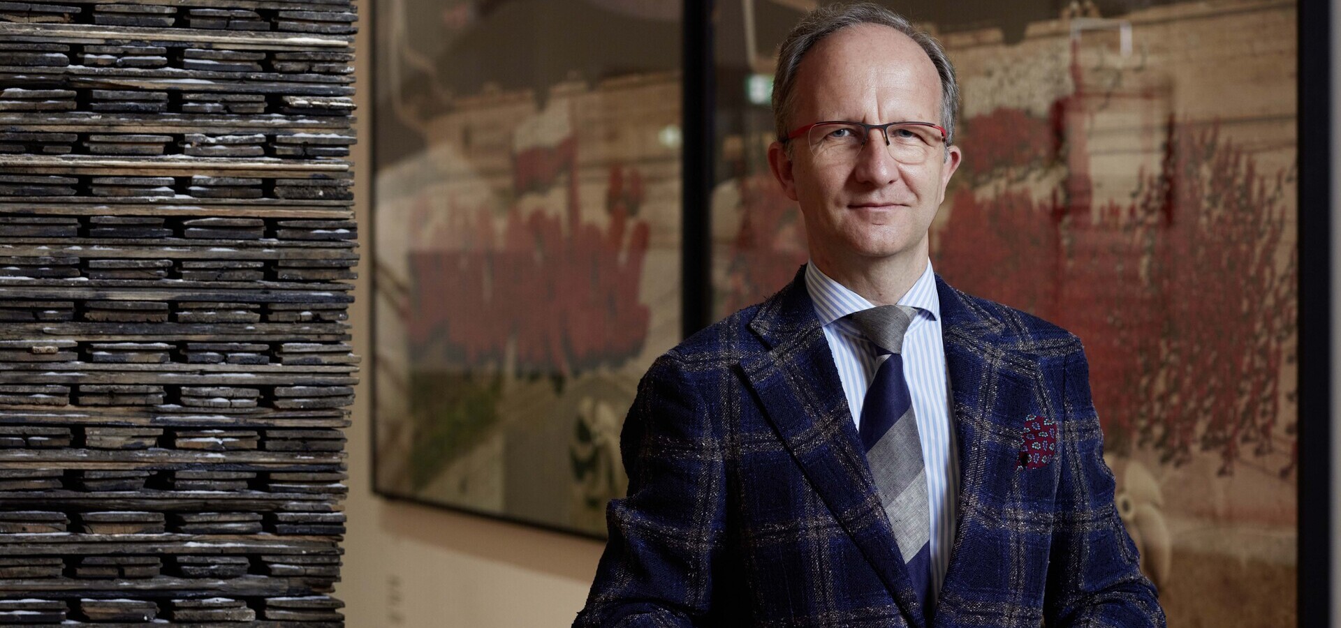 Mężczyzna w okularach stojący po prawej stronie kadru, ubrany w granatową kraciastą marynarkę, jasnoniebieską koszulę i szaro-granatowy krawat, z lewej wysoki pionowy stos ciemnych, płaskich płytek, a w tle rozmyte obrazy z czerwonymi plamami.