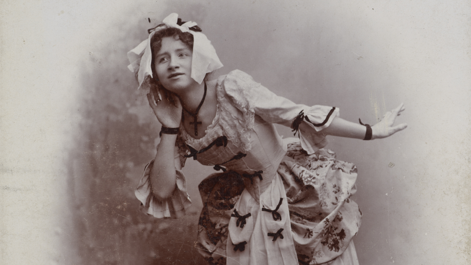 Sepiowa, postarzana fotografia kobiety w historycznym kostiumie, pochylonej do przodu z jedną ręką przy uchu i drugą wyciągniętą w bok, w białym czepcu z kokardami, koronkowej sukni ozdobionej czarnymi kokardkami i naszyjnikiem z krzyżem, na neutralnym tle z widocznymi zadrapaniami.