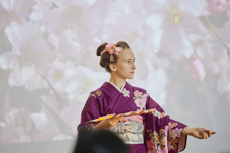 Osoba w profilu na pierwszym planie ubrana w fioletowe kimono z różowo‑złotymi motywami kwiatowymi, z włosami upiętymi w kok ozdobionym różowymi kwiatuszkami, trzymająca zdobiony wachlarz na rozmytym tle dużych jasnoróżowych kwiatów.